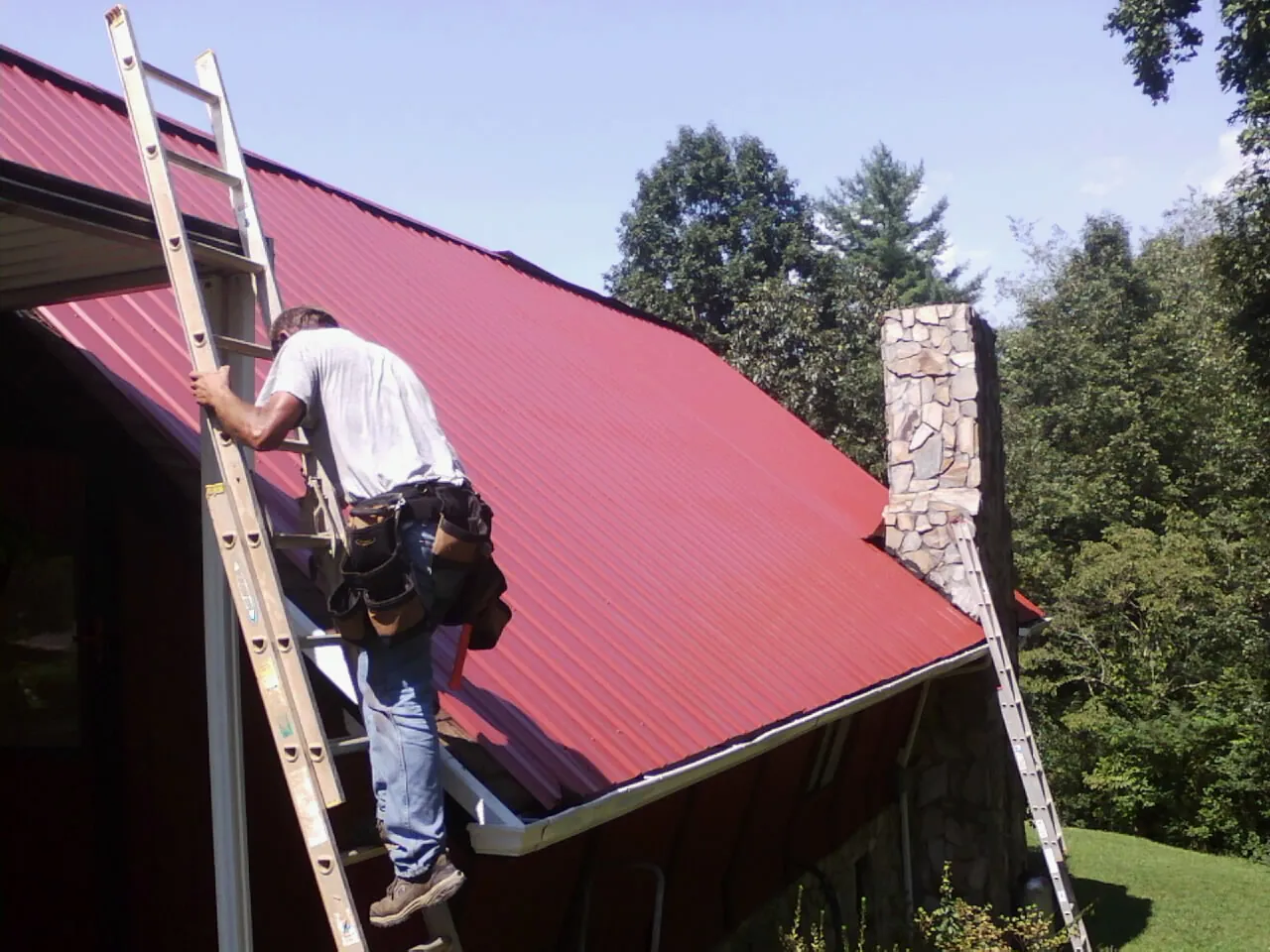 Roofer on a ladder beside a red metal roof and stone chimney — Soffit & Fascia Repair by Roofing Brothers in Florham Park